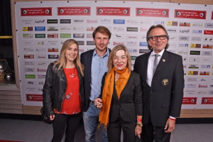 Vier Personen posieren für ein Gruppenfoto vor einer Sponsorwand mit Logos, im Vordergrund mit Anzug- und festlicher Kleidung.