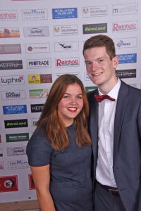 Zwei Personen posieren lächelnd für ein Event-Foto vor einer mit Logos bedruckten Sponsorenwand; der Hintergrund dient als Programmhinweis.