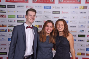 Drei Personen posieren lächelnd vor einer Sponsorwand, Mann im Anzug mit Fliege und zwei Frauen in Abendkleidern bei einem Empfang.