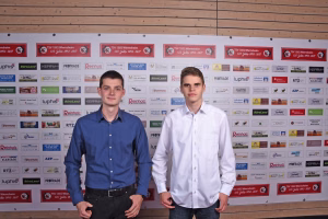 Zwei Jugendliche stehen nebeneinander vor einer Sponsorwand und halten Posen für ein Gruppenfoto bei einer Veranstaltung.