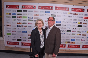 Zwei Personen stehen vor einer Sponsorwand, halten für ein Pressefoto die Position; formelle Kleidung, neutraler Gesichtsausdruck.