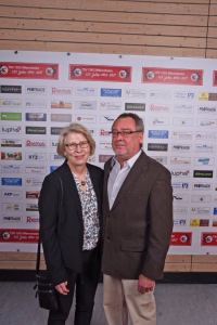 Zwei Personen stehen gemeinsam vor einer Sponsorwand und posieren für ein Foto bei einer Veranstaltung.