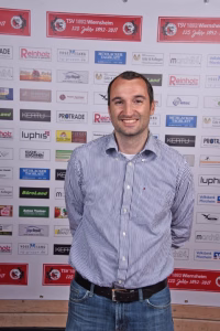 Lächelnder Mann steht vor einer Wand mit vielen Sponsorlogos, als Porträtaufnahme für ein Event/Interview.