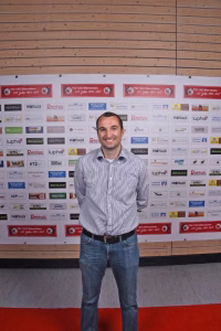 Mann steht vor einer Sponsorwand mit vielen Aufklebern; lächelt in die Kamera, trägt Hemd und Jeans, wirkt wie Veranstaltungsfoto.