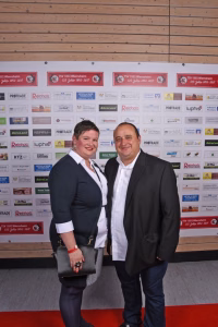 Zwei Personen posieren nebeneinander vor einer Sponsorwand, im Anzug und Businesskleidung, für ein Veranstaltungsfoto.