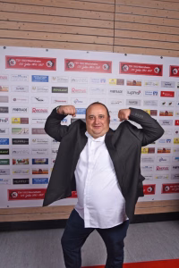 Mann im Anzug auf Event-Lounge, hebt beide Fäuste in Siegerpose vor Sponsorenwand mit Logos und Jubiläumsplaketten.