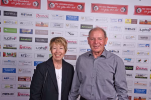 Zwei Personen lächeln vor einer Sponsorenwand mit vielen Firmenlogos; für ein Foto bei einem Event oder Presseauftritt.