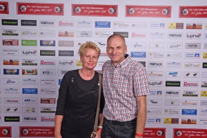 Zwei Personen posieren lächelnd vor einer Sponsorwand mit Logos und Schriftzügen; Aufnahme für ein Event- oder Pressefoto.