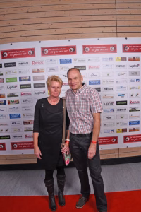 Paar steht gemeinsam vor Sponsorwand und hält beim Event eine Pose auf rotem Teppich, beide blicken in die Kamera.