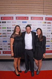 Drei Personen posieren auf rotem Teppich vor einer Sponsorwand; sie präsentieren sich für ein Eventfoto.
