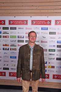 Mann steht frontal vor einer Sponsorwand mit vielen Logos und Vereinsbannern, hält lächelnd die Position für ein Foto bei Veranstaltung.
