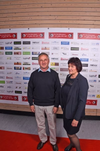 Zwei Personen stehen nebeneinander vor einer Sponsorwand und werden für ein offizielles Gruppenfoto bei einer Veranstaltung präsentiert.