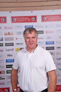 Mann in weißem Poloshirt vor einer Sponsorwand mit Logos, blickt in die Kamera; Portrait für einen Vereins- oder Pressekontext.