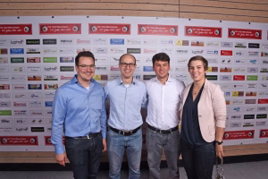 Vier Personen posieren nebeneinander vor einer Sponsorwand; das Bild zeigt Teamfoto-Inszenierung bei einer Veranstaltung mit Logo-Hintergrund.