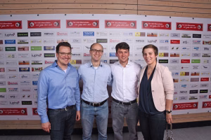 Vier Personen posieren vor einer Sponsorwand, lächeln in die Kamera; Firmenlogos und Textfelder bilden den Hintergrund für ein Gruppenfoto.