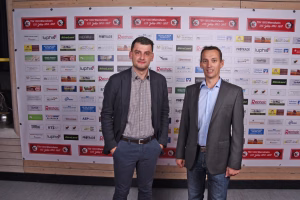 Zwei Männer in Anzügen stehen vor einer Sponsorenwand, lächeln in die Kamera; Hintergrund dient als Event- und Förderhinweis.