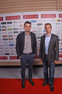 Zwei Personen posieren vor einer Sponsorwand mit Logo-Reihen; formales Outfit, gemeinsames Foto bei Veranstaltung.