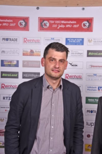 Porträt einer Person vor einer Sponsorenwand mit Logos; Hintergrund dient als Kontext für ein Event oder Presseauftritt.