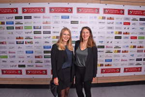 Zwei Frauen posieren lächelnd vor einer Sponsorenwand mit vielen Logos und Plaketten bei einer Veranstaltung.