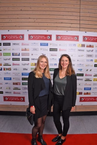 Zwei Frauen stehen nebeneinander auf rotem Teppich vor einer sponsorbedruckten Wand; sie posieren für ein Veranstaltungsfoto.