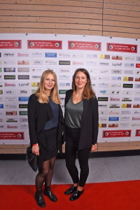 Zwei Frauen posieren vor einer Sponsorwand bei einer Veranstaltung, stehend auf rotem Teppich und mit neutralem Blick in die Kamera.