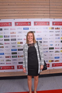 Lächelnde Frau steht vor einer Fotowand mit vielen Vereinslogos, posiert für ein Presse- oder Eventfoto.