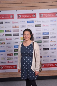 Frau steht lächelnd vor einer Sponsor- und Vereinswand; auf dem Hintergrund sind Logos und Jubiläumsbanner, als Fotohintergrund für einen Auftritt.
