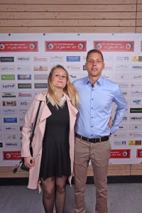 Zwei Personen posieren nebeneinander vor einer Sponsorwand; die Frau trägt Mantel und Kleid, der Mann Hemd und Krawatte, Hände in Hüften bzw. an Seite.