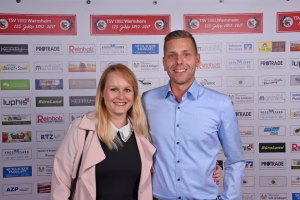 Zwei Personen posieren lächelnd vor einer Wand mit vielen Firmenlogos und Vereinsbanner; wirkt wie ein offizielles Teamfoto.