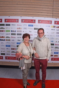 Zwei Personen mit Medaille auf rotem Teppich vor Wand voller Vereinslogos und Sponsoraufkleber, lächelnd für ein Erinnerungsfoto.