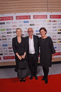 Drei Personen posieren auf rotem Teppich vor einer Sponsorwand, lächeln in die Kamera und markieren einen offiziellen Anlass.