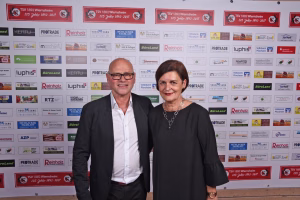 Zwei Personen stehen vor einer Sponsorwand, lächeln in die Kamera; formelles Event-Foto zur Darstellung der Teilnehmenden.