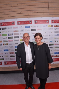 Zwei Personen stehen nebeneinander auf rotem Teppich vor einer Sponsor-Wand mit Logos und Jubiläums-Aufdrucken.