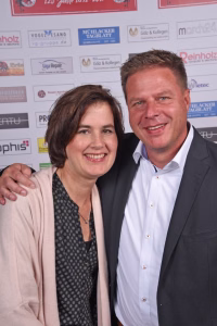 Zwei Personen posieren lächelnd nebeneinander bei einer Pressewand, um ein gemeinsames Erinnerungsfoto bei einem Event zu zeigen.