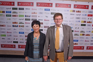 Zwei Personen posieren nebeneinander vor einer Wand mit zahlreichen Sponsorlogos, aufgenommen bei einer offiziellen Veranstaltung.