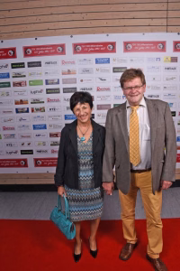 Zwei Personen stehen lächelnd auf rotem Teppich vor einer Sponsorwand mit Logos, im Rahmen eines offiziellen Anlasses.