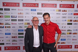 Zwei Männer posieren nebeneinander vor einer Wand mit Sponsor-Logos, einer lacht und hält den anderen am Arm, beide lächeln in die Kamera.