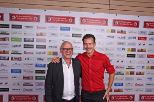 Zwei Männer posieren lächelnd vor einer Sponsorwand, einer im roten Hemd und einer im dunklen Jackett; Arm über die Schulter.