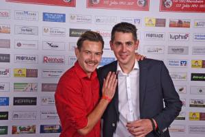 Zwei Männer posieren vor einer Sponsorwand, einer hält die Hand an die Schulter des anderen; freundlicher Gruß bei einer Veranstaltung.