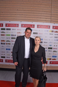 Zwei Personen in Anzügen posieren auf einem Event vor einer Sponsorwand; sie stehen für ein offizielles Foto im Hintergrund.