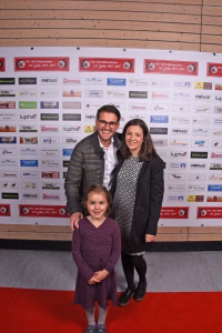 Zwei Erwachsene und ein Kind posieren vor einer Sponsorenwand; die Familie steht Schulter an Schulter für ein Gruppenfoto bei einer Veranstaltung.