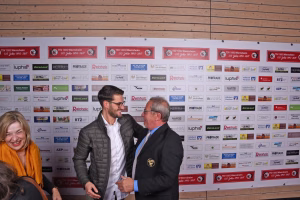 Zwei Männer im Gespräch vor einer Sponsorwand mit vielen Firmenlogos; eine Person lächelt, die andere spricht, im Hintergrund Holzrahmen.