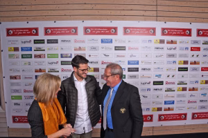 Zwei Männer und eine Frau im Gespräch vor einer Sponsorwand, Hände zur Begrüßung, Anzug und Freizeitkleidung im Hintergrund.