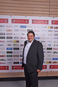Mann im Anzug lächelnd vor einer Sponsorwand mit vielen Logos, aufgenommen bei einer Veranstaltung.