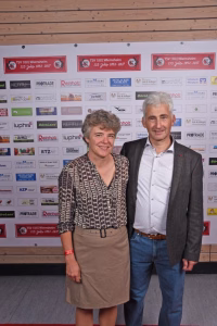 Zwei Personen stehen nebeneinander vor einer Sponsorwand, lächeln in die Kamera; Schirmfoto eines Veranstaltungsfotos.