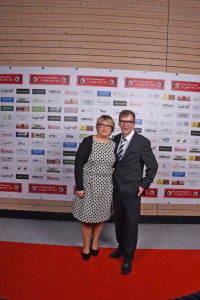 Zwei Personen stehen auf rotem Teppich vor einer Sponsorwand mit zahlreichen Logos, wirken wie bei einem Event- oder Fotoauftritt.