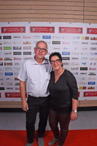 Zwei Personen stehen gemeinsam vor einer Sponsorwand und halten für ein Event-Foto die Pose, im Hintergrund sind Logos und Festplaketten.