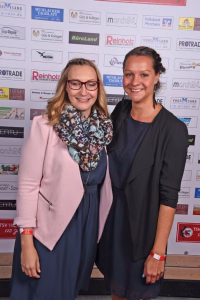 Zwei Frauen lächeln und posieren vor einer Wand mit vielen Logo- und Sponsorenanzeigen, wirken wie bei einer Veranstaltung.