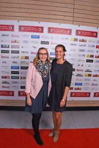 Zwei Frauen stehen nebeneinander vor einer Sponsorwand und dem roten Teppich, lächeln in die Kamera.