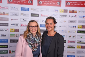 Zwei lächelnde Personen vor einer Wand mit vielen Logos und Namensschildern, als Porträt bei einer Veranstaltung.